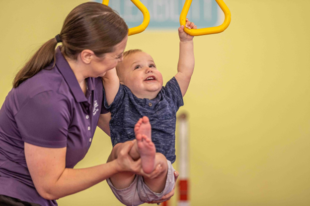 A coach is helping a toddler at the little gym doing hanging bars activities